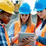 A group of young, diverse construction professionals, including men and women, engaged in a discussion on a construction site. They are collaboratively reviewing digital blueprints on a tablet, representing the integration of technology and diversity in the industry.