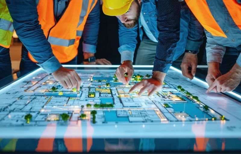 A diverse team of architects, engineers, and site workers collaborating around an interactive digital table, showcasing building plans and fostering inclusive design practices.