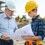 "A senior construction manager and a young site supervisor review detailed building plans on a bustling construction site, emphasizing mentorship and leadership development within the construction industry."