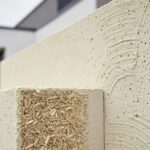 Close-up of a modern wall section with hempcrete texture in light beige tones, with visible hemp hurd shiv and binder materials in the foreground and a building exterior in soft focus behind it.