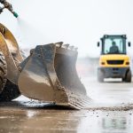 Industrial pressure washer spraying water to remove mud from an excavator bucket and nearby site equipment on a construction yard.